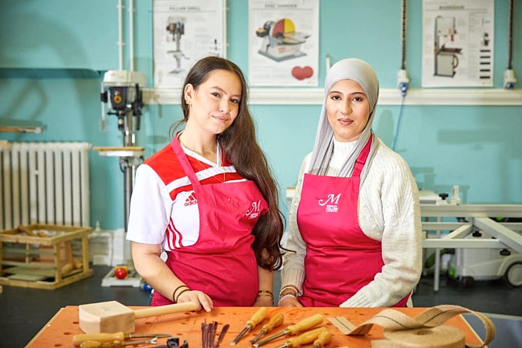 Two young female students one with hijab standing at woodwork desk
